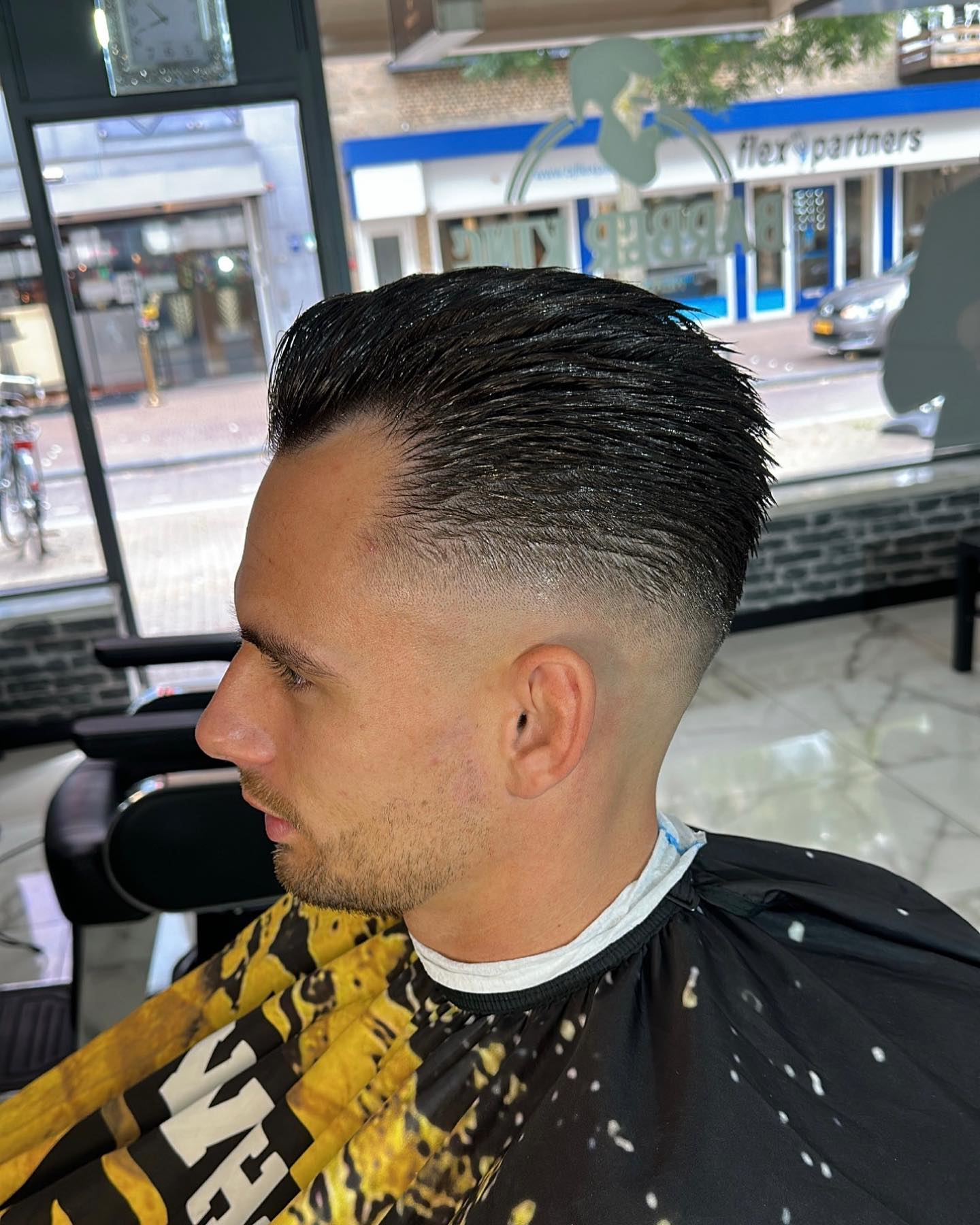 Klant met achterovergekamd haar en een hoge fade, bij het raam van de zaak